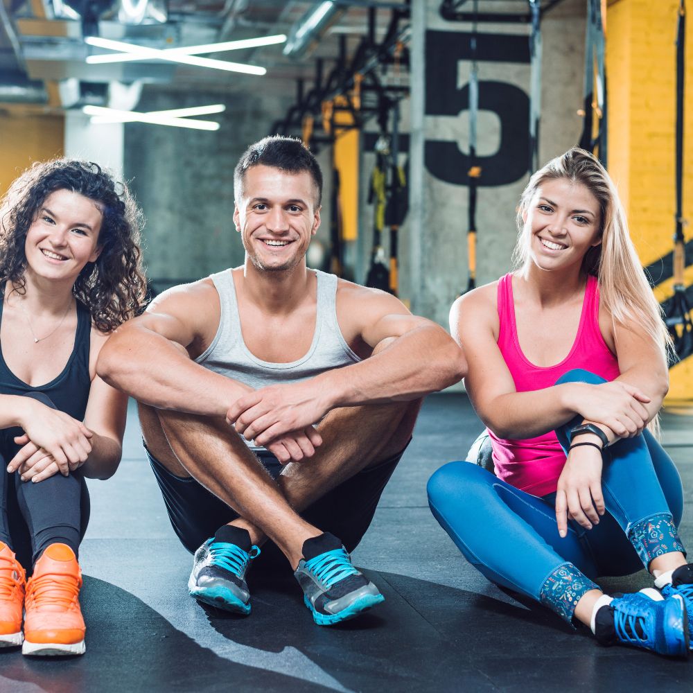 Three fit people sitting on the gym floor in a fitness center, wearing athletic clothing and sneakers. The background shows gym equipment and a large number 5 on a wall.